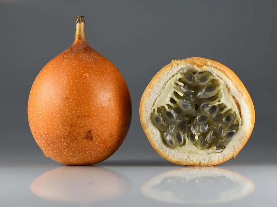 Granadilla entera de cáscara anaranjada con puntos blancos junto a una mitad cortada que muestra la pulpa gris translúcida con semillas negras envueltas en arilos