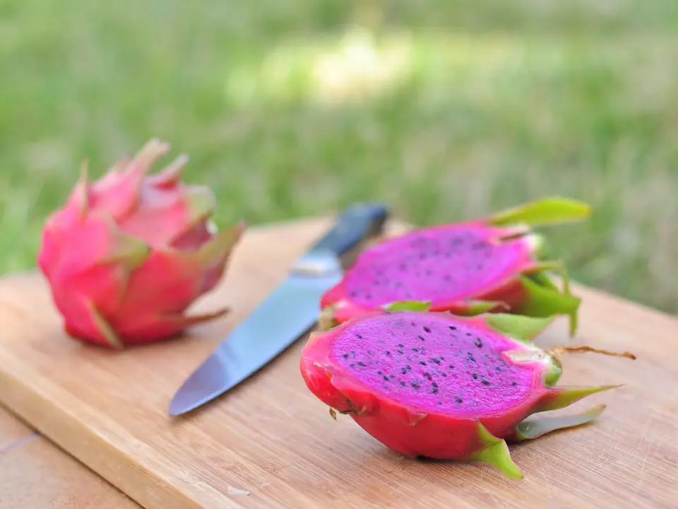 Dos mitades de pitahaya roja con pulpa magenta y semillas negras sobre una tabla de madera, con una fruta entera y un cuchillo detrás, al aire libre.