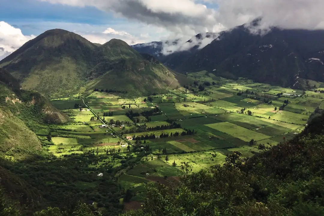 Valle andino cultivado con parcelas verdes de distintos tonos, iluminadas por el sol y rodeadas de montañas cubiertas de nubes
